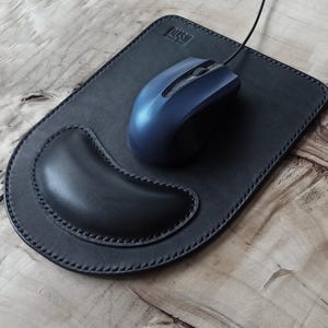 以下が含まれることがあります： 手首用のクッションと青いワイヤレスマウスが付いた黒い革製のマウスパッド。マウスパッドには「LUSH」という文字が刻印されています。