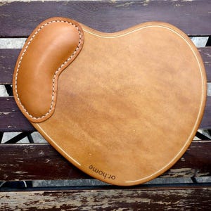Può includere: Tappetino per mouse a forma di cuore in pelle marrone chiaro con poggiapolsi integrato. Il poggiapolsi è un rettangolo arrotondato, cucito al cuscinetto principale con filo bianco. Bordo cucito e testo "or.home".