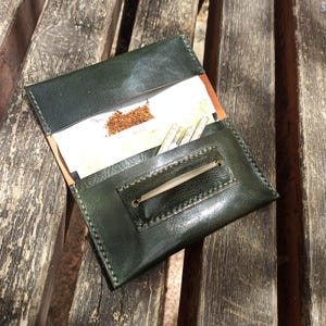 Puede incluir: Bolsa de tabaco de cuero verde abierta, con tabaco y cigarrillos enrollados. La bolsa está abierta, mostrando el contenido, y se encuentra sobre una superficie de madera envejecida. La costura es visible y el cuero tiene un ligero brillo.