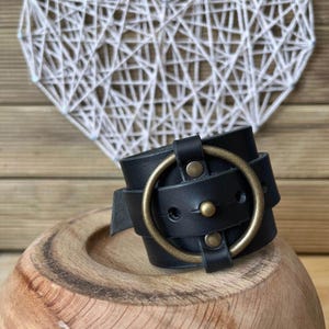 Op de afbeelding: Zwarte leren manchetarmband met een grote messingkleurige ring en klinknagels. De armband is om een houten kom gewikkeld. De achtergrond is een wit hartvormig draadkunstwerk.