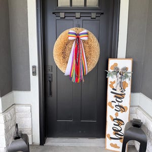 May include: A front door with a round, tan wreath adorned with a rainbow ribbon and colorful streamers. A wooden sign with the text "hey y'all" and cow print accents stands to the right. Black lanterns flank the entrance.