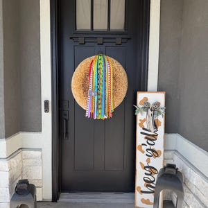 May include: A round, textured wreath with colorful ribbons hangs on a black front door. A wooden sign with the words "hey y'all" and a leopard print design stands to the right of the door. Two black lanterns flank the door.