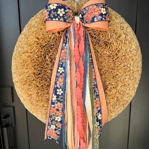 May include: A round, textured wreath with a large bow and flowing ribbons. The bow features a navy blue floral pattern with peach accents. Ribbons in various colors, including peach, blue, and cream, cascade down. The wreath is hanging on a dark gray door.