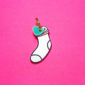 Op de afbeelding: Een witte sokvormige sleutelhanger met zwarte contouren en een rode rand. Een kleine, turquoise hartvormige bedel is met een goudkleurige ring aan de bovenkant bevestigd. De achtergrond is felroze.