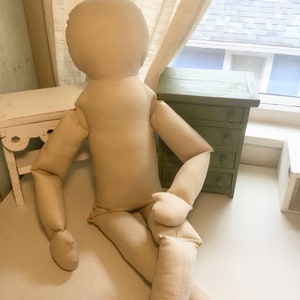 May include: A large, beige fabric doll with a rounded head, arms, and legs, posed in a seated position. The doll is made of a soft, neutral-colored material. It is positioned against a white surface, with a window and furniture in the background.