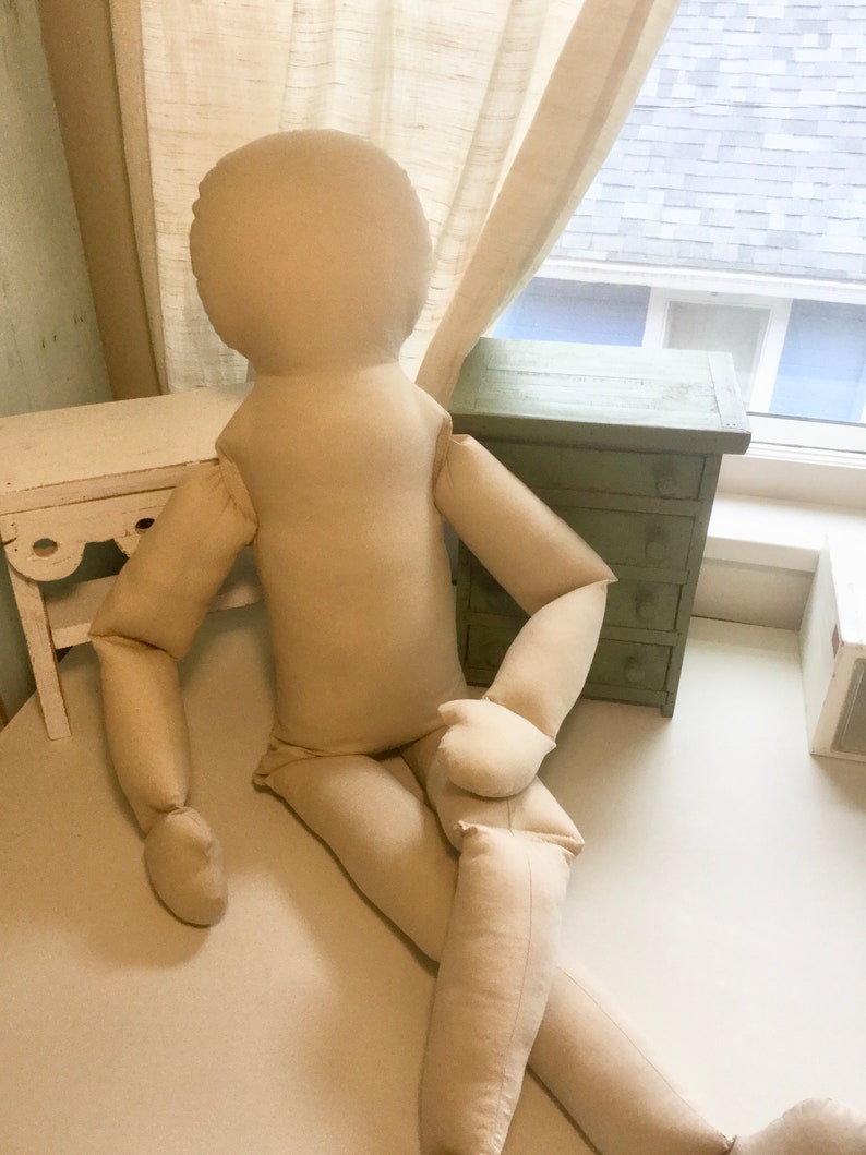 May include: A large, beige fabric doll with a rounded head, arms, and legs, posed in a seated position. The doll is made of a soft, neutral-colored material. It is positioned against a white surface, with a window and furniture in the background.