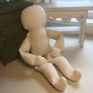 May include: A white fabric doll with a simple design, sitting on a white surface. The doll has a round head, no facial features, and limbs that are segmented.