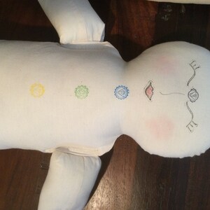 May include: A white fabric doll with embroidered chakra symbols in red, yellow, green, and blue. The doll's face has drawn features, including pink cheeks and a small mouth. The doll is lying on a dark wooden surface.