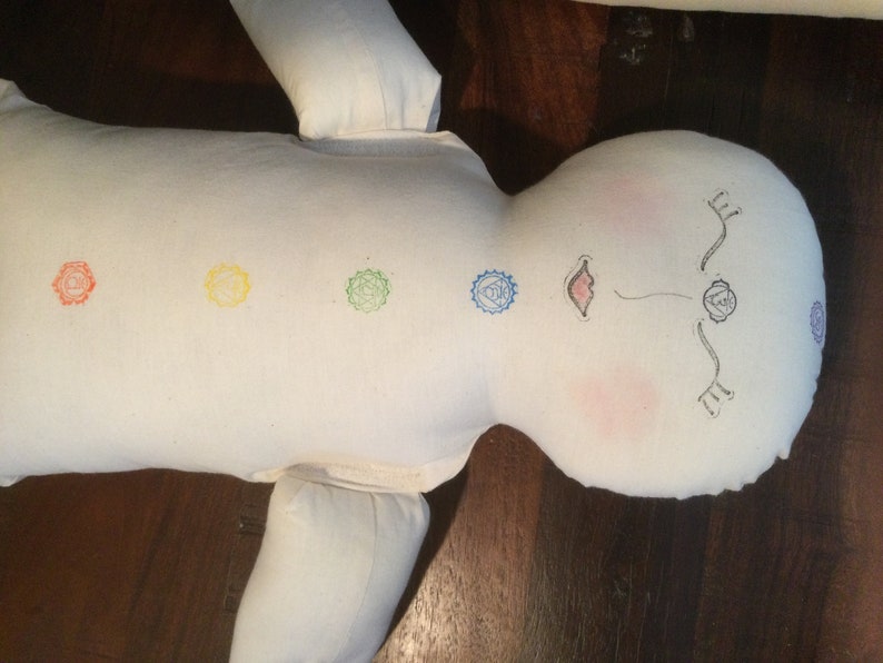 May include: A white fabric doll with embroidered chakra symbols in red, yellow, green, and blue. The doll's face has drawn features, including pink cheeks and a small mouth. The doll is lying on a dark wooden surface.