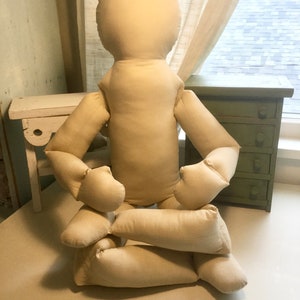 May include: A beige fabric doll with a rounded head, arms, and legs, in a seated position. The doll's limbs are bent, and it appears to be made of a soft, stuffed material. The background includes a small green cabinet and a white bench.