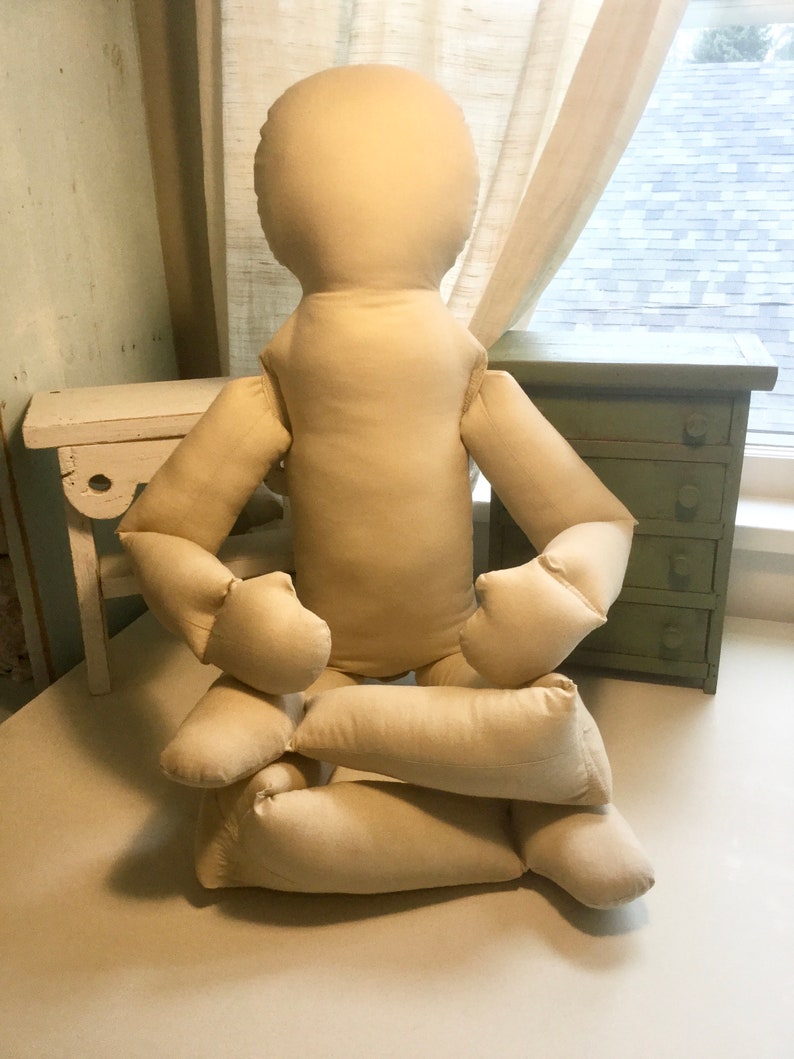 May include: A beige fabric doll with a rounded head, arms, and legs, in a seated position. The doll's limbs are bent, and it appears to be made of a soft, stuffed material. The background includes a small green cabinet and a white bench.