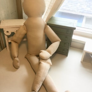 May include: A large, beige fabric doll with a rounded head, arms, and legs. The doll is posed in a seated position with one leg crossed over the other. The doll is made of a soft, unadorned fabric.