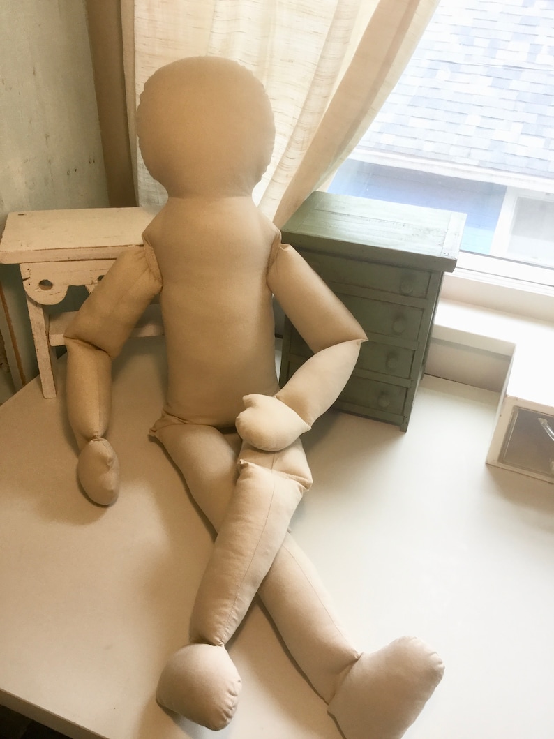May include: A large, beige fabric doll with a rounded head, arms, and legs. The doll is posed in a seated position with one leg crossed over the other. The doll is made of a soft, unadorned fabric.