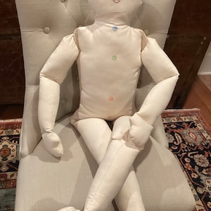 May include: A white fabric doll with a human-like form, sitting in a chair. The doll has a simple embroidered face with closed eyes and a pink mouth. There are colorful circles embroidered on the doll's torso, representing chakras.