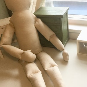 May include: A large, beige fabric doll with a rounded head, arms, and legs, lying on a white surface. The doll is positioned near a small, green wooden cabinet with three drawers. The scene is lit by natural light.