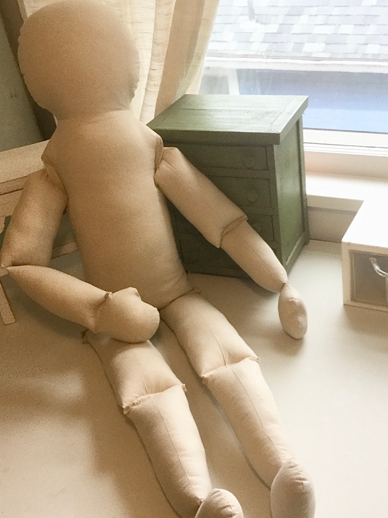 May include: A large, beige fabric doll with a rounded head, arms, and legs, lying on a white surface. The doll is positioned near a small, green wooden cabinet with three drawers. The scene is lit by natural light.