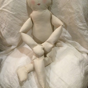 May include: A white fabric doll with a simple painted face, closed eyes, and a pink mouth. The doll has long limbs and is sitting with its legs crossed.