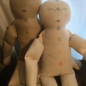 May include: Two beige fabric dolls with embroidered facial features and chakra symbols. The dolls have closed eyes, rosy cheeks, and red lips. Each doll has a series of embroidered circles down the center of the torso, representing the chakras.
