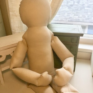May include: A large, beige fabric doll with a rounded head, arms, and legs. The doll is seated with its arms resting on its legs. The doll is made of a soft, unadorned fabric, and is positioned in front of a window and a small green cabinet.