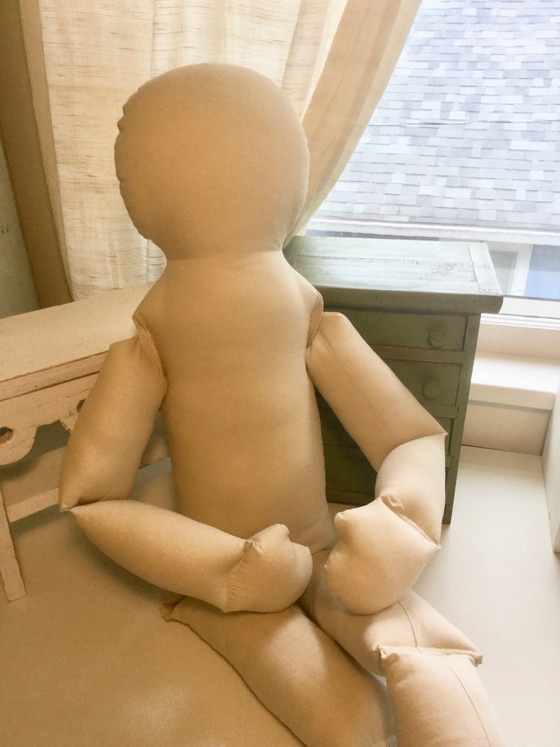 May include: A large, beige fabric doll with a rounded head, arms, and legs. The doll is seated with its arms resting on its legs. The doll is made of a soft, unadorned fabric, and is positioned in front of a window and a small green cabinet.