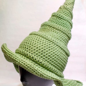 May include: A green crocheted witch hat with a tall, pointed crown and a wide brim. The hat is on a black mannequin head.