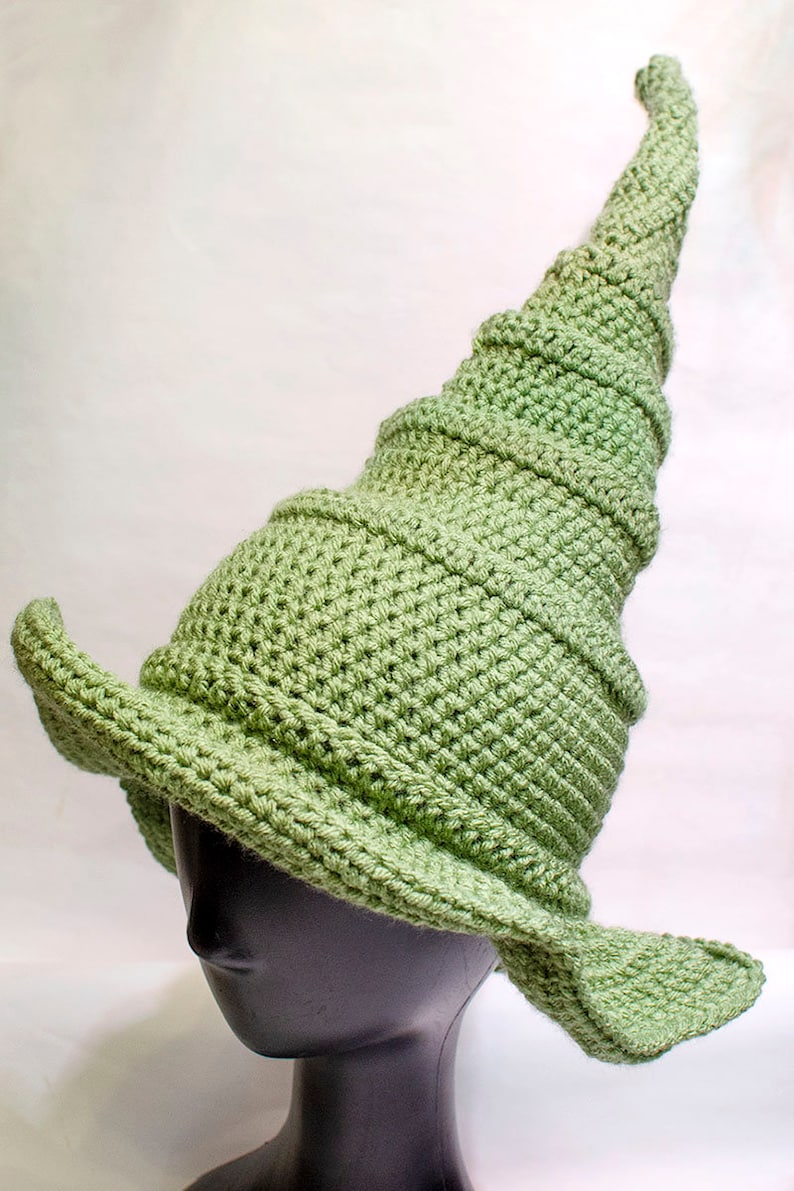 May include: A green crocheted witch hat with a tall, pointed crown and a wide brim. The hat is on a black mannequin head.