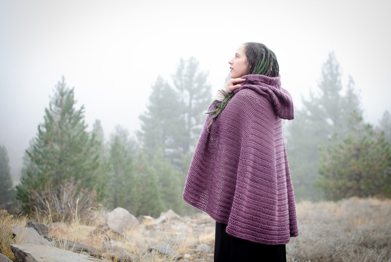 Pu&ograve; includere: Una donna indossa un poncho di uncinetto viola con cappuccio in un ambiente boschivo nebbioso.