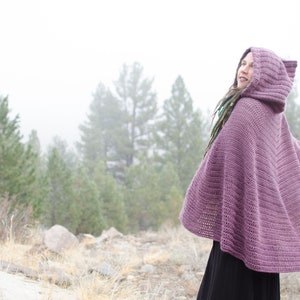 Pu&ograve; includere: Un mantello di crochet viola con cappuccio e un motivo a punto testurizzato. Il mantello &egrave; indossato da una persona in piedi in una foresta nebbiosa.