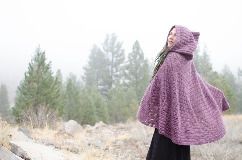 Pu&ograve; includere: Un mantello di crochet viola con cappuccio e un motivo a punto testurizzato. Il mantello &egrave; indossato da una persona in piedi in una foresta nebbiosa.