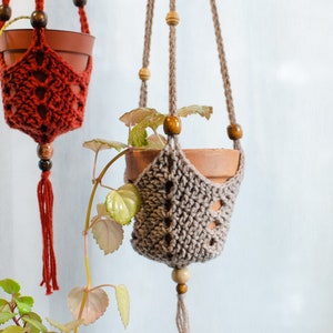 Può includere: Due vasi sospesi per piante realizzati in macramè all'uncinetto con perline di legno. Il vaso a sinistra è rosso e il vaso a destra è beige.
