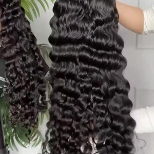 May include: A long, black, wavy wig with a deep wave pattern. The wig is held up, showcasing the length and volume of the hair. The hair appears shiny and healthy, with a natural-looking texture. The wig is designed for a full head of hair.