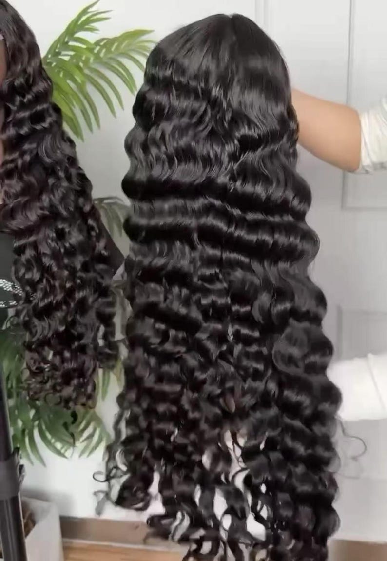 May include: A long, black, wavy wig with a deep wave pattern. The wig is held up, showcasing the length and volume of the hair. The hair appears shiny and healthy, with a natural-looking texture. The wig is designed for a full head of hair.