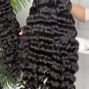 May include: Two black, wavy wigs are displayed. The wigs have long, voluminous, curly hair. The hair is a deep black color and has a natural shine. The wigs are held up, showcasing the full length and texture of the hair. The background is a neutral color.