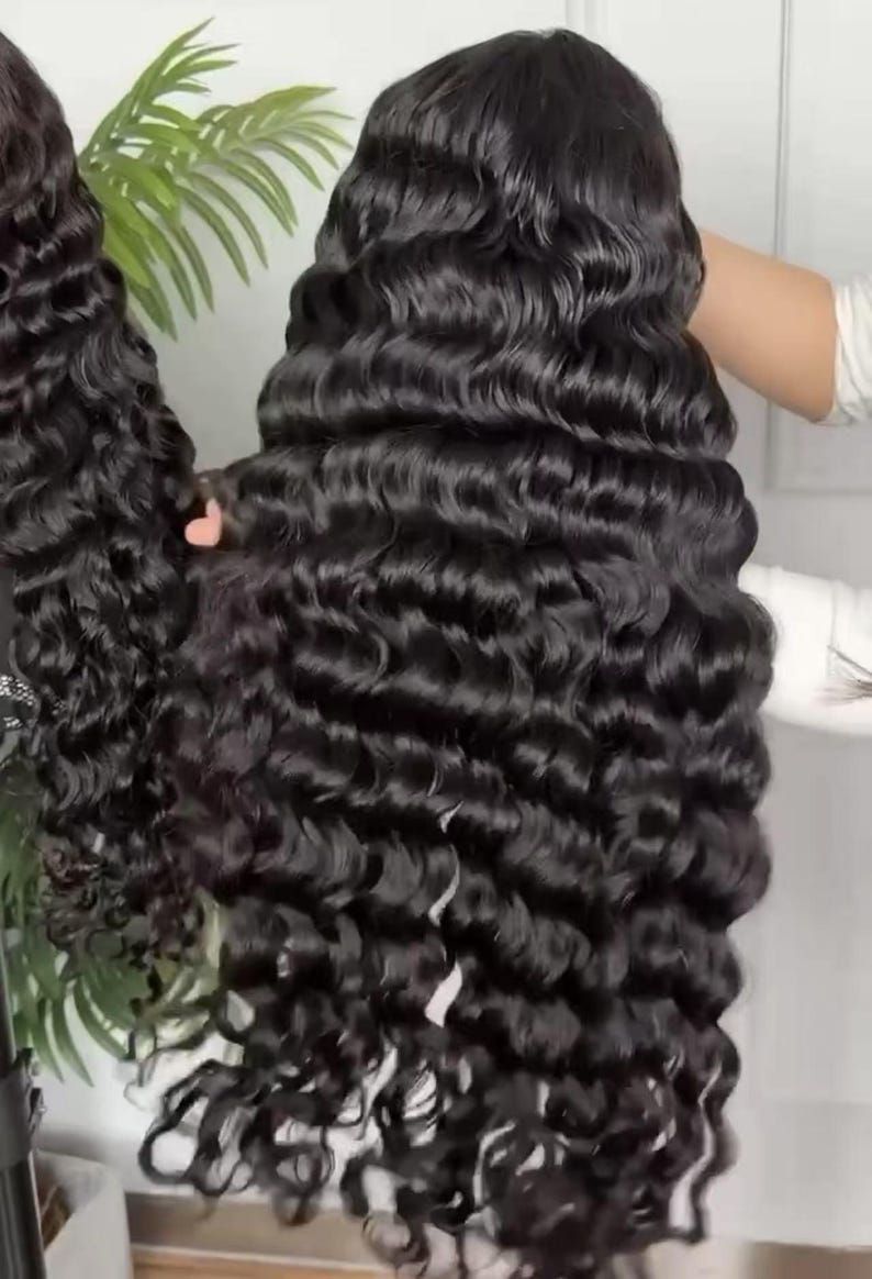 May include: Two black, wavy wigs are displayed. The wigs have long, voluminous, curly hair. The hair is a deep black color and has a natural shine. The wigs are held up, showcasing the full length and texture of the hair. The background is a neutral color.