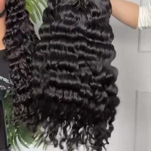 May include: A long, black, wavy wig with a deep wave pattern. The wig is held up by a person's hand, showcasing the voluminous curls and the natural-looking hairline. The hair appears shiny and well-styled, suitable for various occasions.