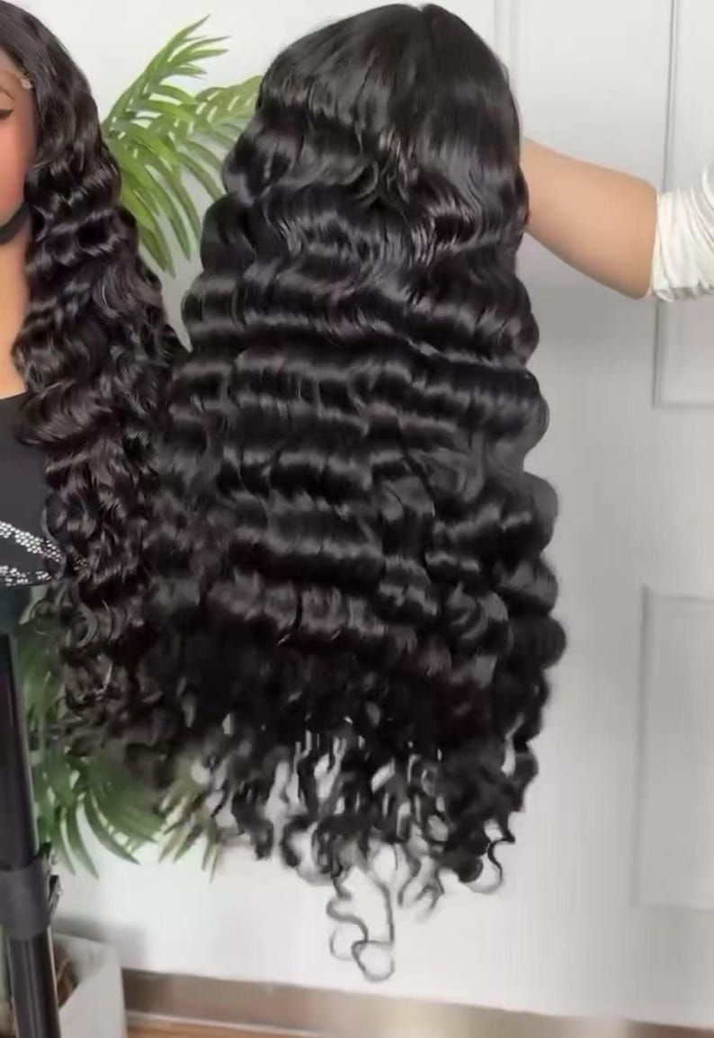 May include: A long, black, wavy wig with a deep wave pattern. The wig is held up by a person's hand, showcasing the voluminous curls and the natural-looking hairline. The hair appears shiny and well-styled, suitable for various occasions.