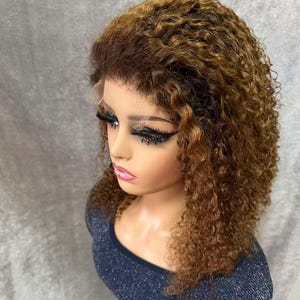 May include: A mannequin head showcasing a curly, brown wig with highlights. The wig has a lace front and is styled in a shoulder-length, voluminous cut. The mannequin is wearing a dark blue, sparkly top.