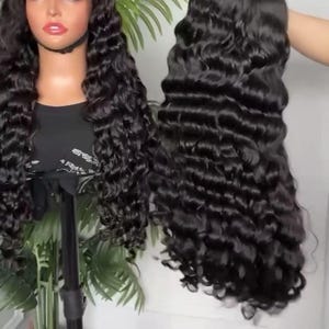 May include: Two black curly wigs are displayed. One is on a mannequin head, and the other is held up by a hand. The wigs have long, wavy, and voluminous hair. The hair is a deep, rich black color, and the curls are well-defined.