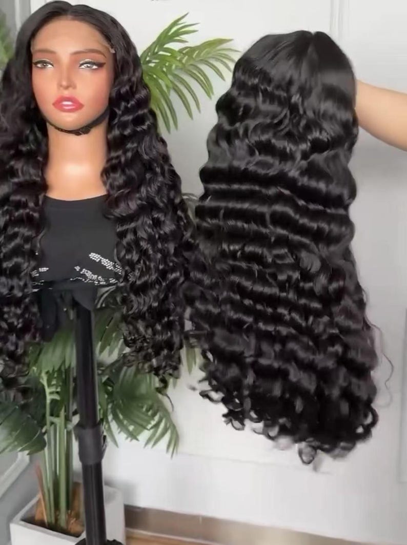 May include: Two black curly wigs are displayed. One is on a mannequin head, and the other is held up by a hand. The wigs have long, wavy, and voluminous hair. The hair is a deep, rich black color, and the curls are well-defined.