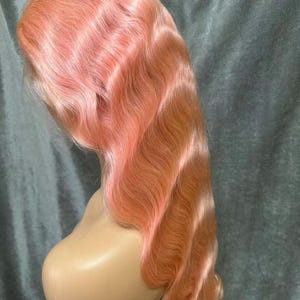 May include: A peach-coloured wig with long, wavy hair. The wig is styled with deep waves and is displayed on a mannequin head. The hair has a gradient effect, transitioning from a lighter peach at the roots to a darker peach at the ends.