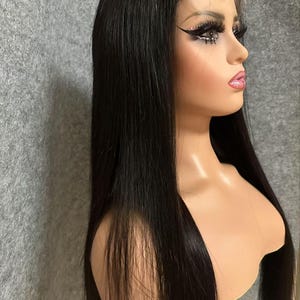 May include: A long, straight black wig with a lace front. The wig is styled with a centre parting and falls to the shoulders.