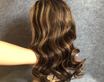 May include: A brown wig with blonde highlights and long, wavy curls. The wig is styled to fall around the shoulders and has a natural-looking hairline. The hair is a mix of dark brown and golden blonde.