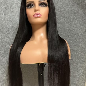 May include: A long, straight black human hair wig on a mannequin head. The wig is styled in a centre part and has a natural, sleek look.