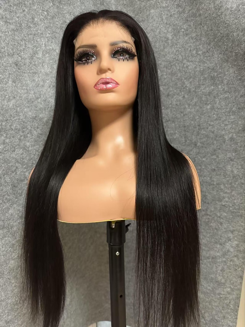 May include: A long, straight black human hair wig on a mannequin head. The wig is styled in a centre part and has a natural, sleek look.