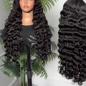 May include: A black, long, wavy wig on a mannequin head, displayed on a black stand. The wig has a lace front and is styled with voluminous, cascading curls. A second wig of the same style is shown to the right, against a white background.