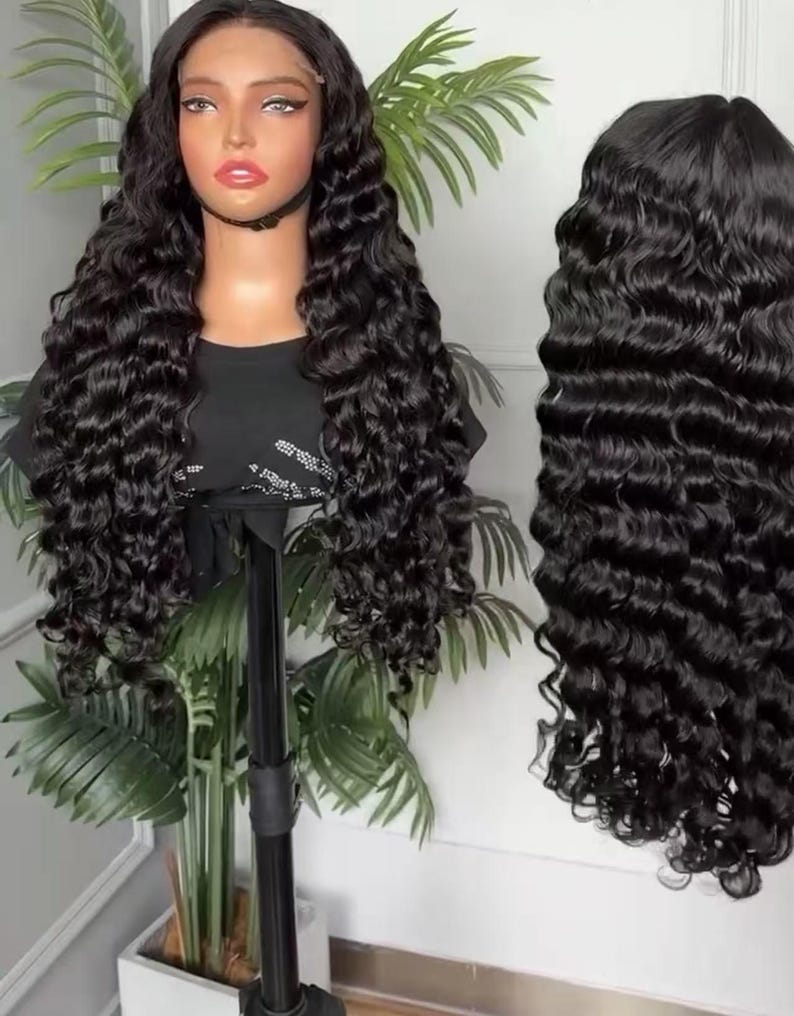 May include: A black, long, wavy wig on a mannequin head, displayed on a black stand. The wig has a lace front and is styled with voluminous, cascading curls. A second wig of the same style is shown to the right, against a white background.