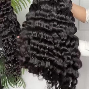 May include: A long, wavy black wig with voluminous curls. The wig is held up, showcasing the texture and style of the hair. The curls are tightly wound and cascade down, creating a full and glamorous look.