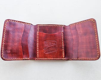 May include: A tri-fold leather wallet in a deep reddish-brown hue with visible tan stitching along the edges. The interior features multiple card slots and a central pocket embossed with the text Bluffland Leather Co.