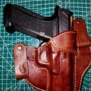 May include: A black handgun rests in a brown leather holster. The holster is stitched with light-colored thread and has a belt loop. The handgun has a textured grip. The brand name "Burkland" is embossed on the holster.