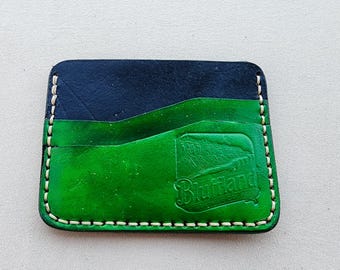 May include: A rectangular leather card holder featuring a two-tone design with a dark navy blue top section and a vibrant green bottom section. The piece is stitched with light-colored thread and embossed with the text Bluffland.